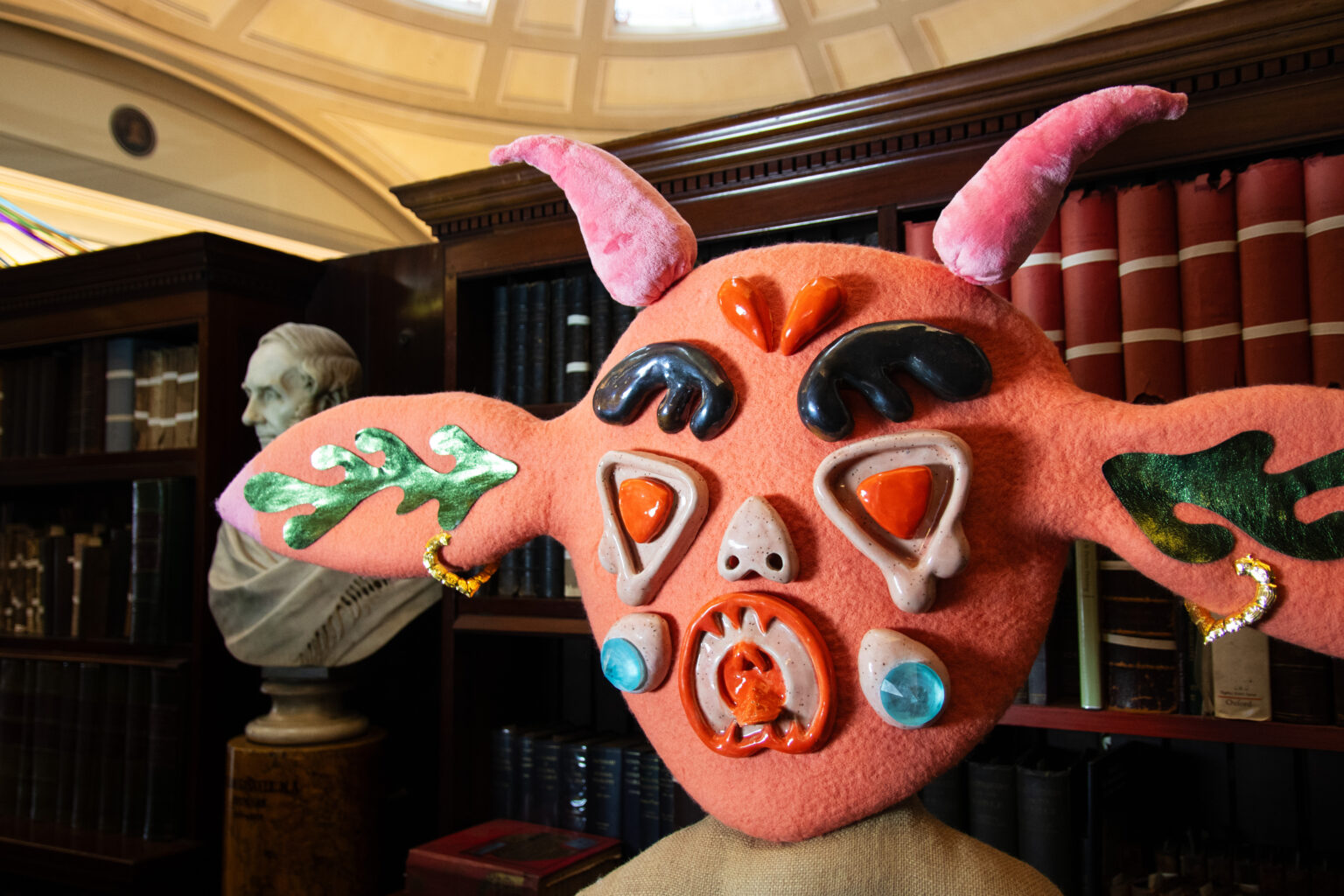 A close up of the textile head of a creature whose felt skin is peach coloured with pink horns atop their head and long ears that protrude right and left, with facial features of colourful plastic or ceramic - large black eyebrows, red eyes, round open mouth and blue gemstone cheeks.