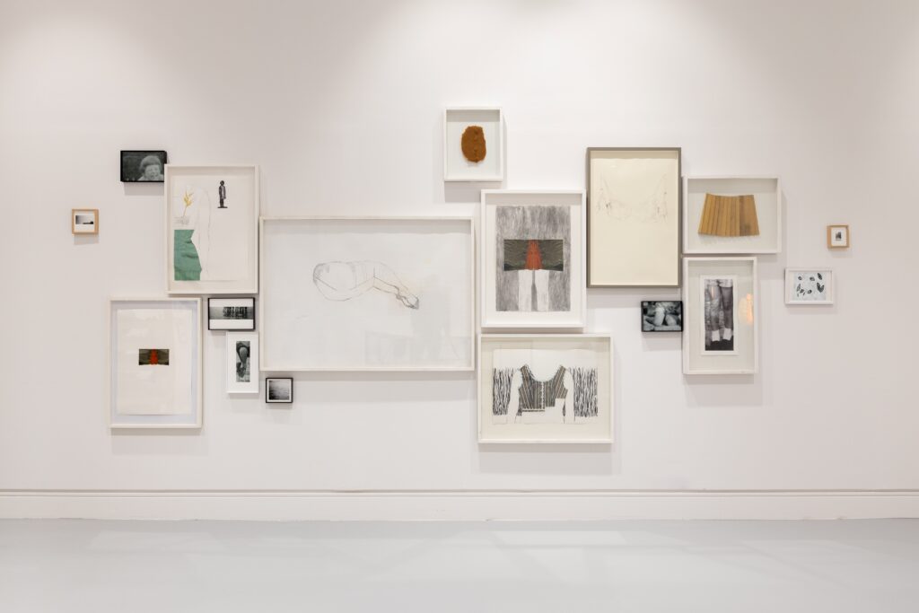 A white wall with an arranged cludter of framed drawings and photographs.