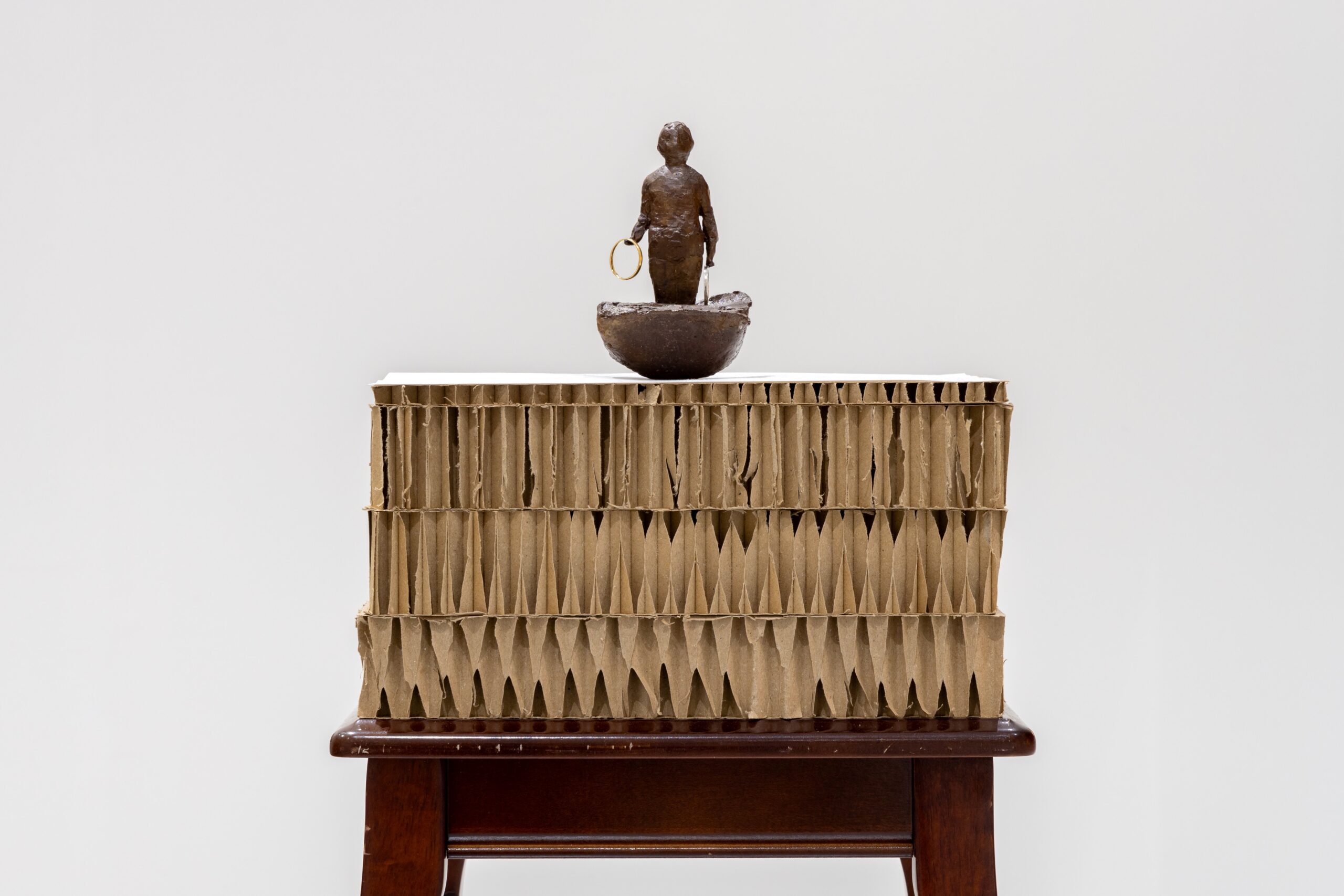 A small bronze sculpture of a girl holding rings in a boat-like form, perched on top of a thick square of cardboard on top of a small, long-legged occasional table.