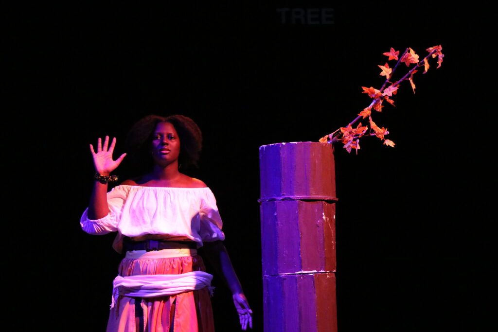 Camara with her right hand raised, paml towards the audience and fingers spread, beside the constructed tree trunk with two branches emerging