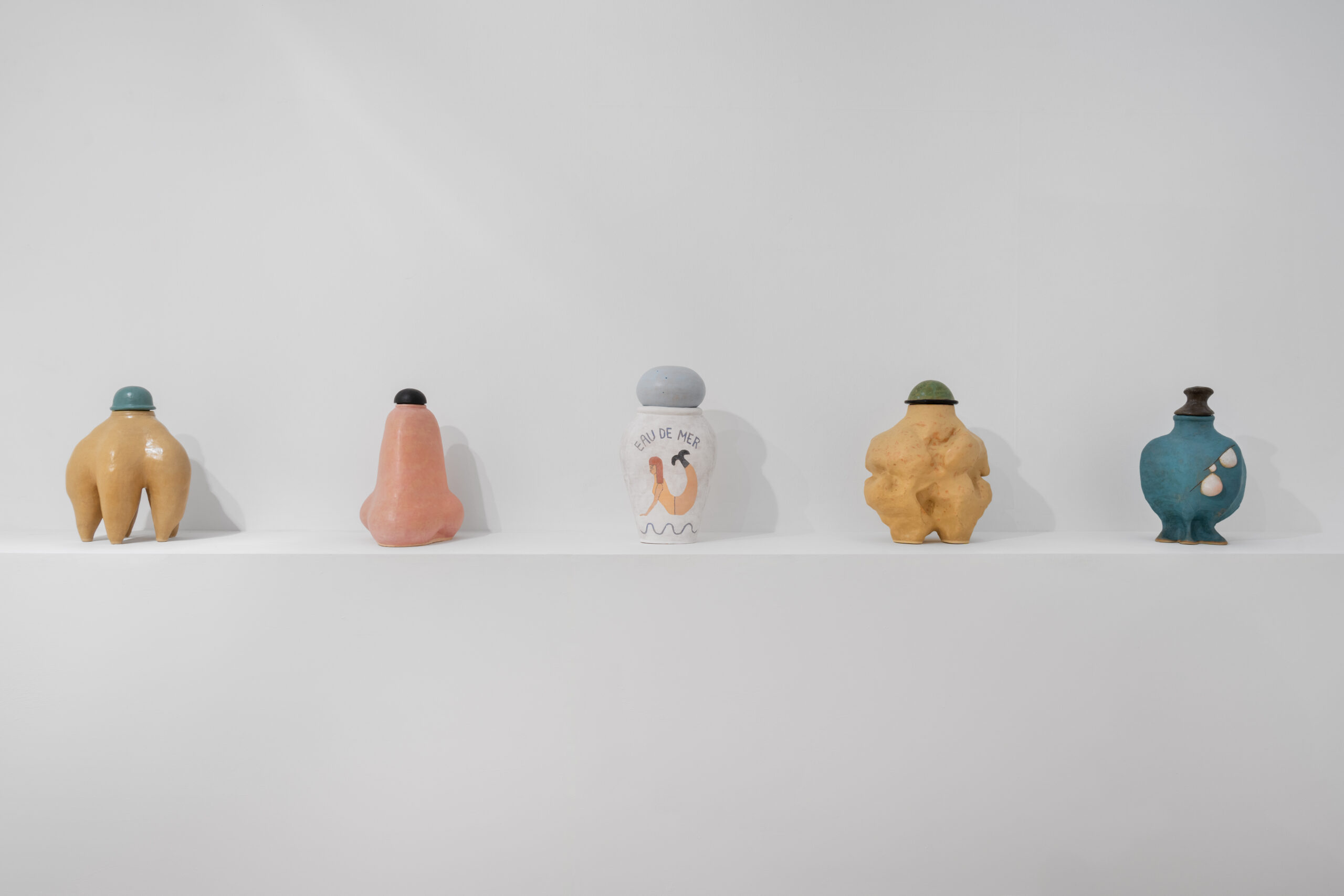 Five little ceramic bottles on a white shelf, from left to white yellow with tentacles, a pink nose, a white bottle with a mermaid on, a squishy venus figure and a blue bottle