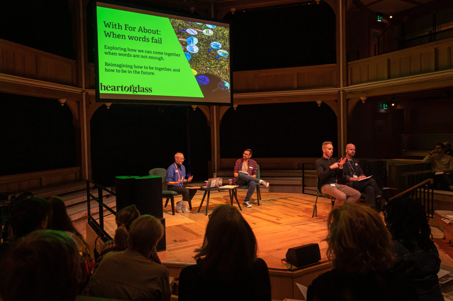 four people sit on stage. Two people are in conversation with one another. The other two people are sign interpreters and translate the conversation facing an audience.