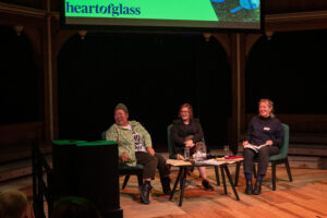 Three people sit together on a stage. Between them is a table on which sit glasses of water and sheets of paper.