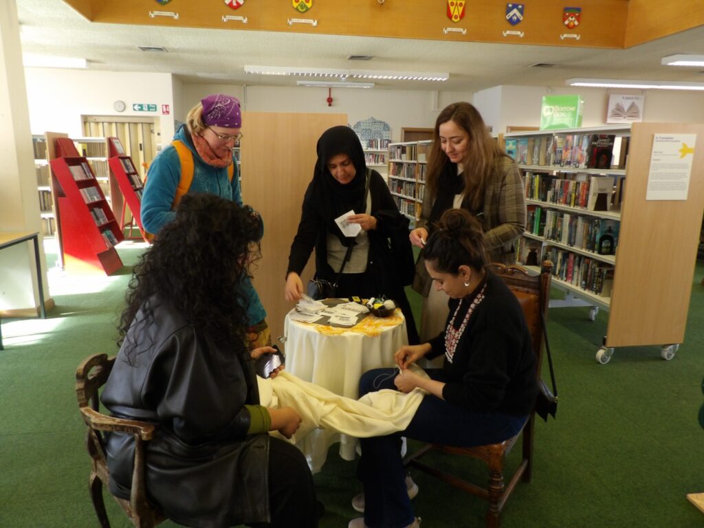 Inside a library room with shelves of books two people sit at a small table, one sewing something onto a piece of fabric the other holds, while three people stand on the other side of the table looking