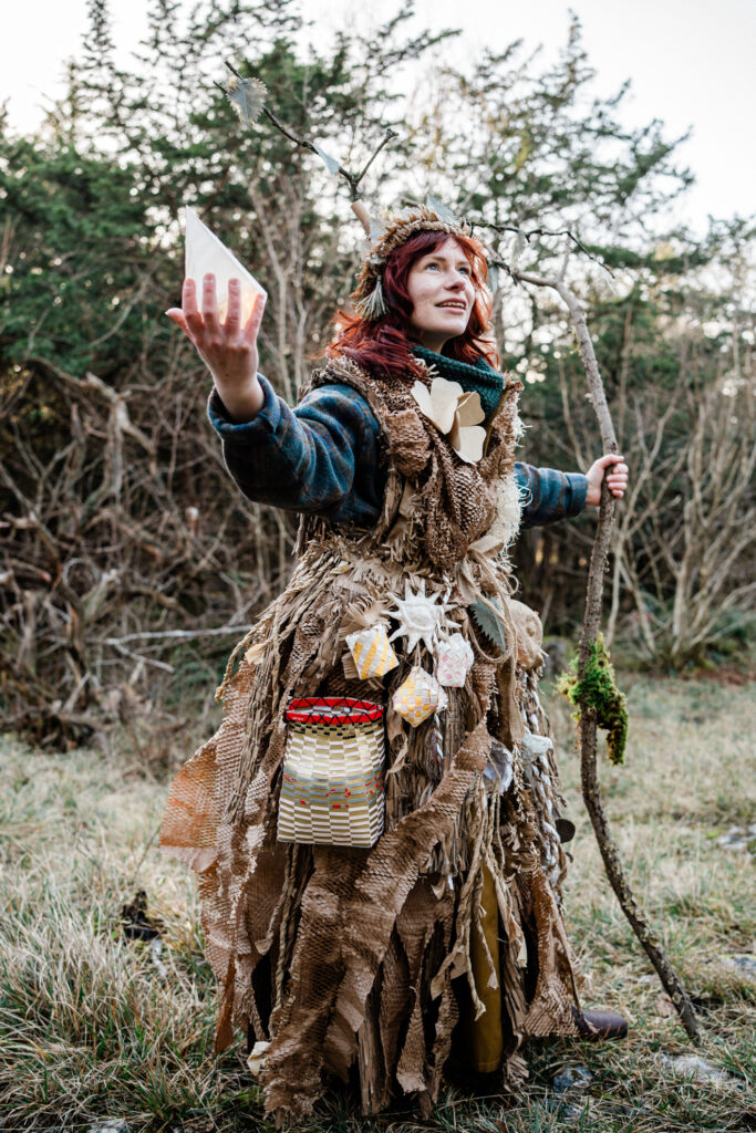 A woman wearing a costume of browns covered with little trinkets and pockets and baskets, carrying a staff