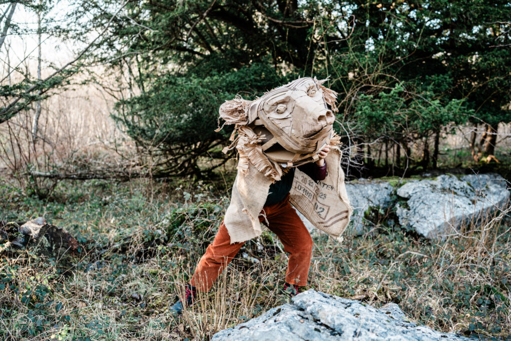 A huge paper mask of a bear over a persons body dancing in the woods, you can see their little human legs sticking out the bottom