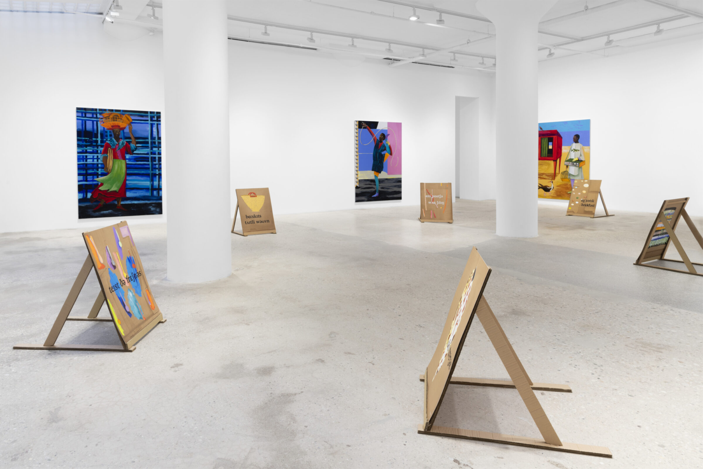 A white walled gallery with three large bright paintings on the walls and cardboard signs propped up on the floor around the space