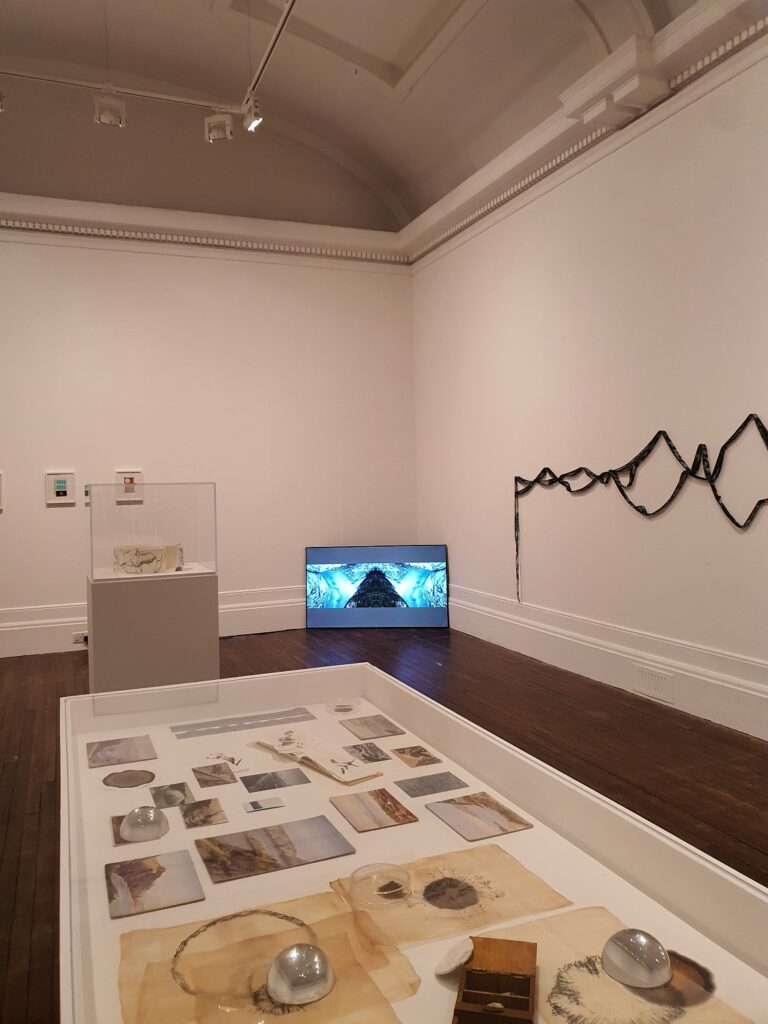 The corner of the gallery including a vitrine with images and objects, a TV screen in the corner and draped black ribbon along one wall