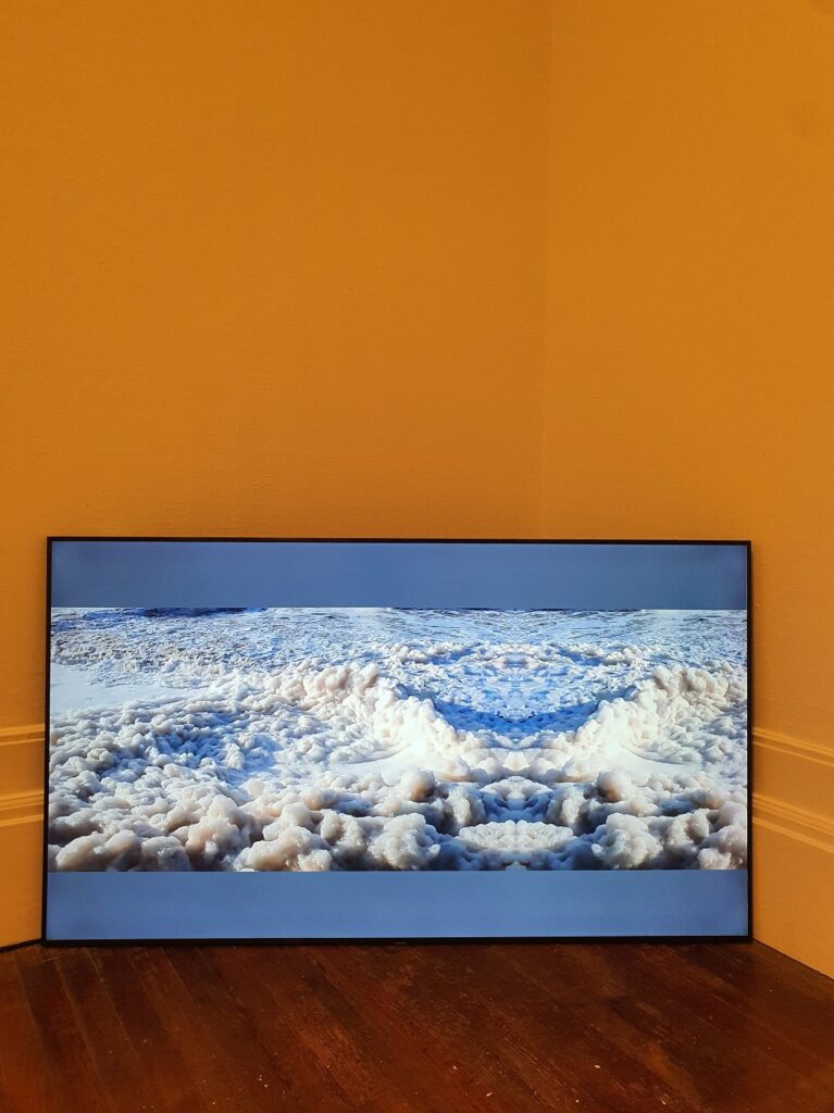 A wide screen at the floor of an orange wall which shows blue footage of rolling sea waves