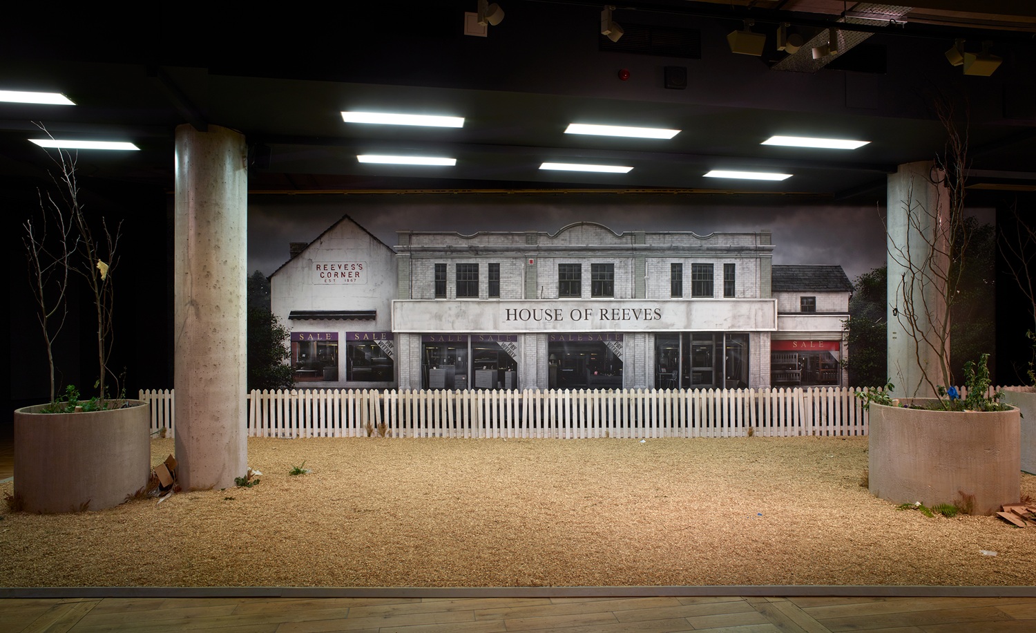The stage set of House of Reeves shop front behind a white picket fence and gravel floor, with concrete pillars and planters with dead trees on either side