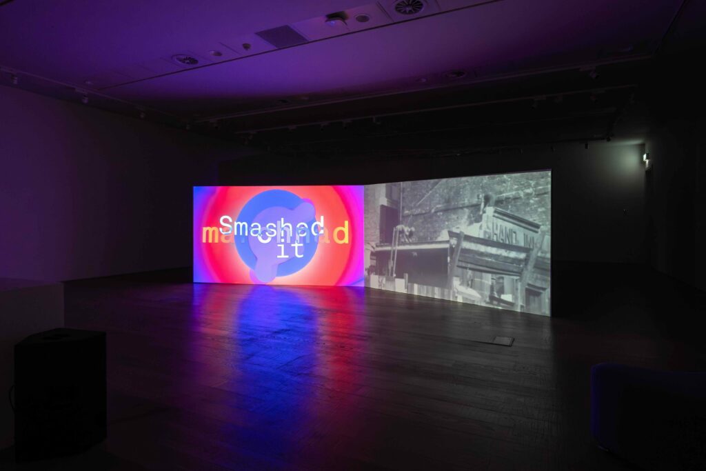 A dual channel film is projected onto a large screen in a gallery. The room is dark, lit only by the film.