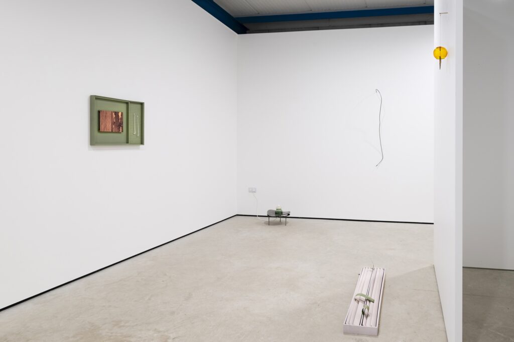 A white wall galley with three small sculptures, a green box on the wall, a small table on the floor and a long white box on the floor