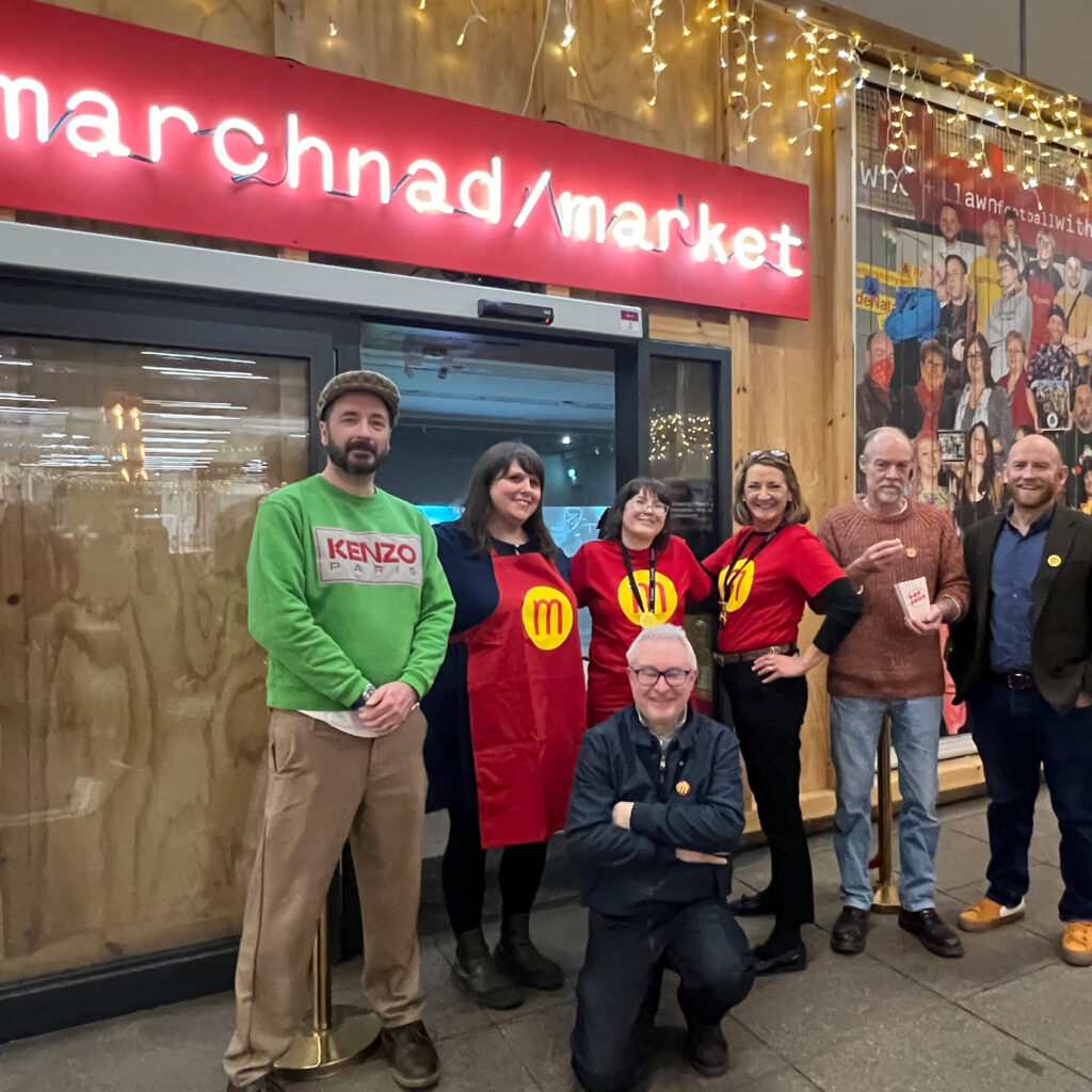 A group of people stand under a neon sign displaying the words 'marchnad/market'.