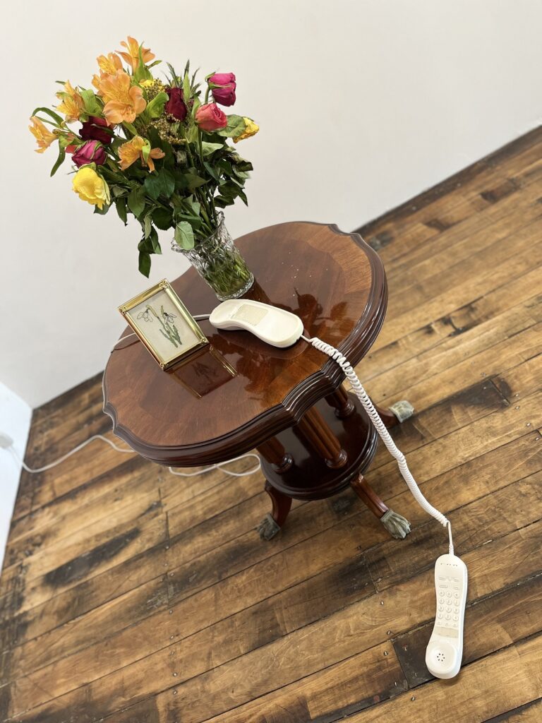 an end table with cable phone off the hook beside a framed image and vase of flowers