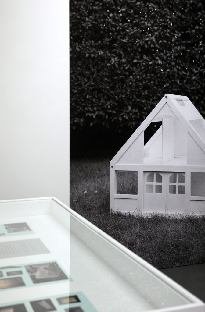 The top of a vitrine with images inside and behind it a huge print of a picture of a white doll's house against a black background