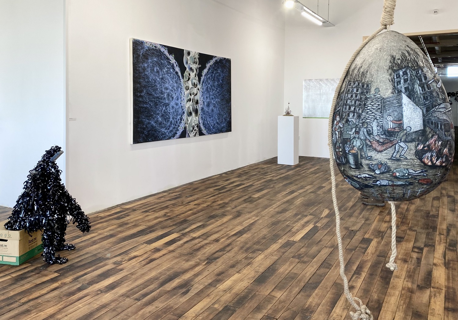 A big egg hanging with images of war on the right, on the left a human shaped figure made of black reels of tape