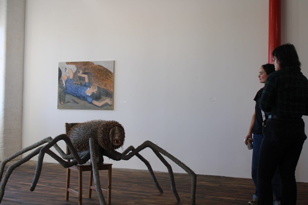 a huge spider resting on a dining chair in front of a painting of a girl being eaten by a bird