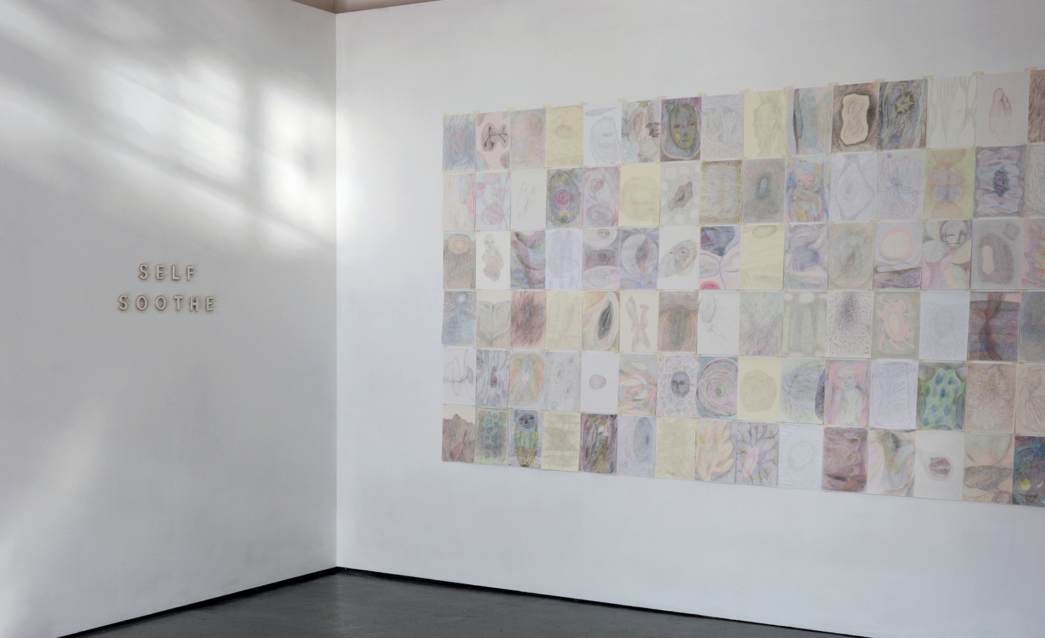 Corner of a white walled room on the left the words SELF SOOTHE in relief and on the right a grid of pale drawings