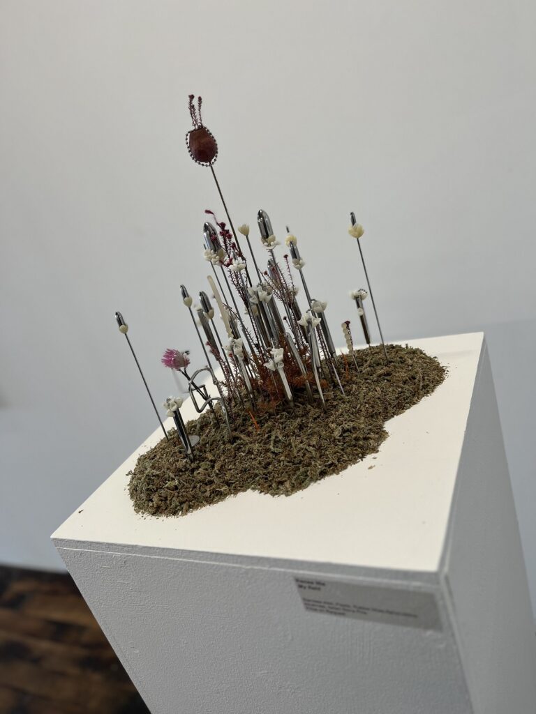 a cityscape of tall metal plants and poles emerge from a heap of leaf mulch on a white plinth