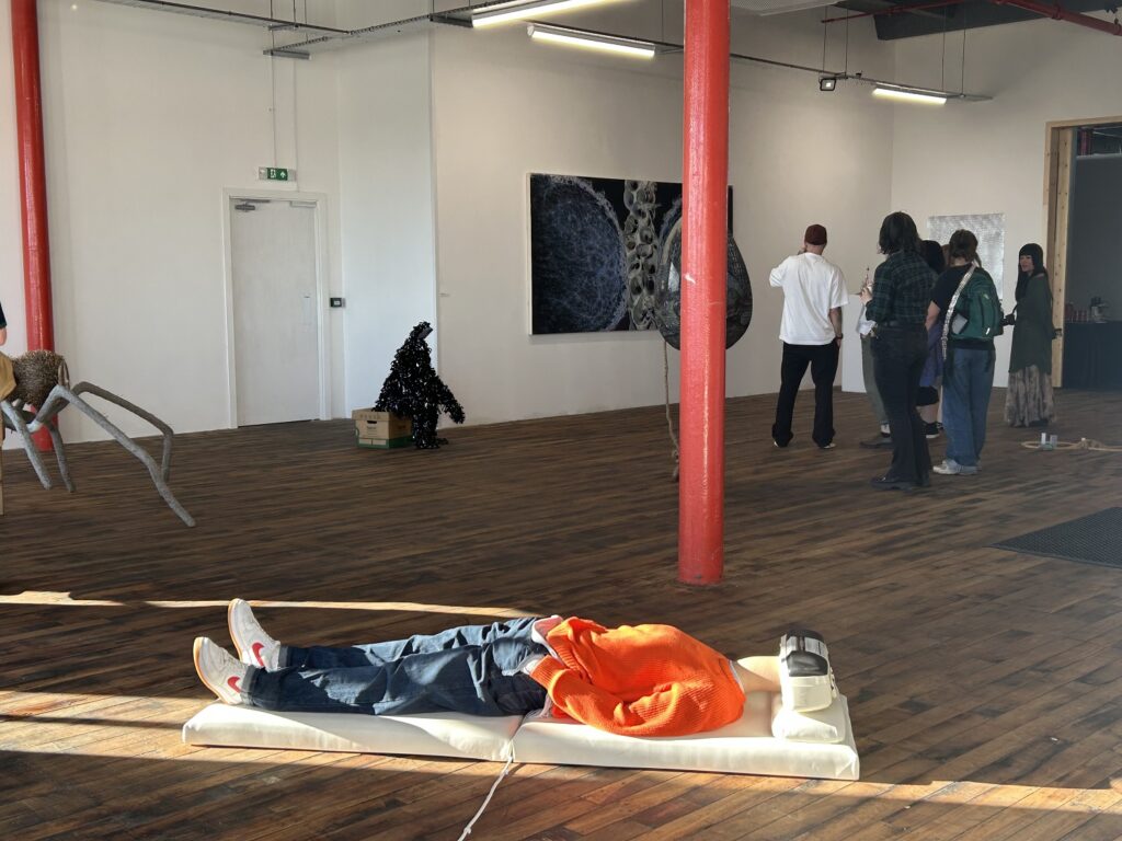 A person in an orange jumper and blue jeans lies on the floor, their head is a tape player