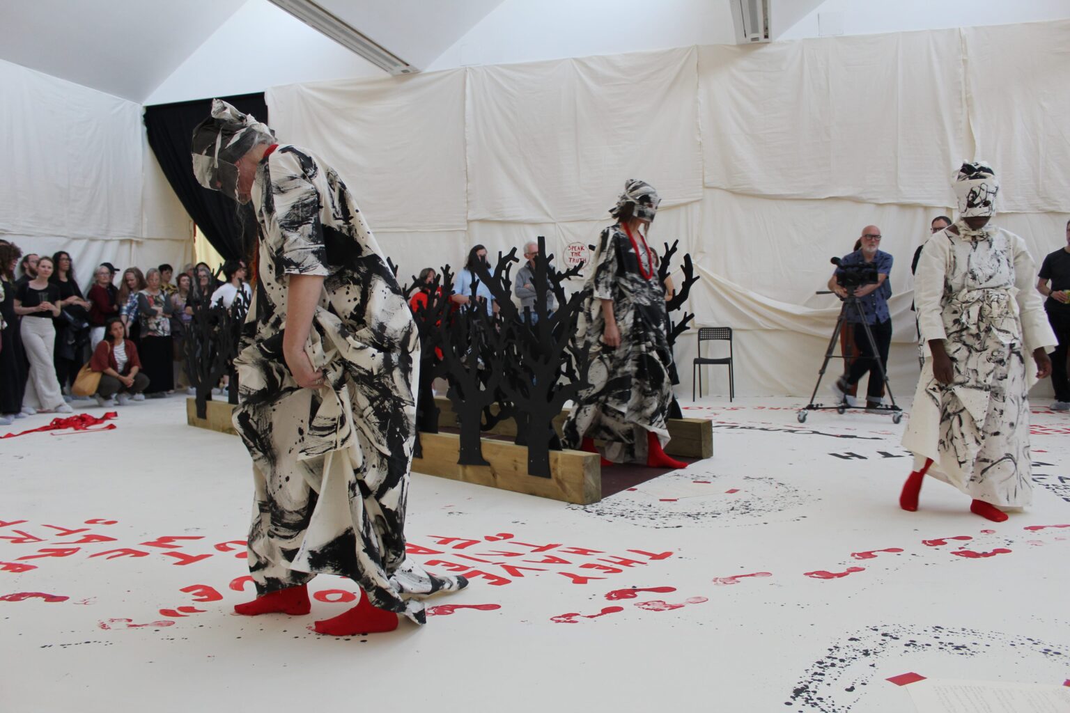 Three performers wearing painted garb traverge a gallery space draped with calico. The floor is white vinyl and has words and fortprinted painted on in black and red. There is an audience watching.