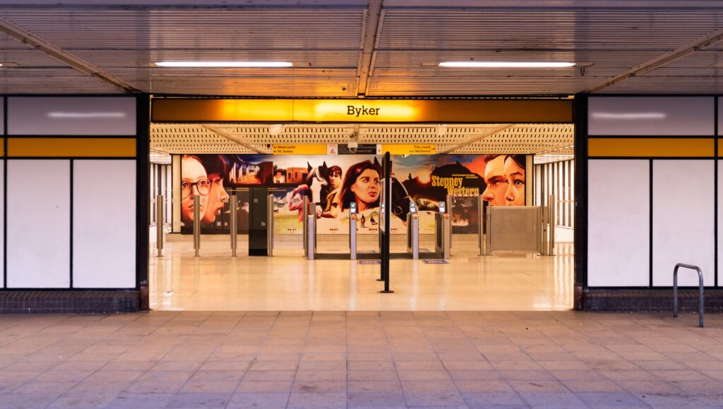 The Tyne and Wear Metro station in Byker, whaite and yellow pannelled. Inside is apainted mural showing young people and horses in the style of an old fashioned film poster.