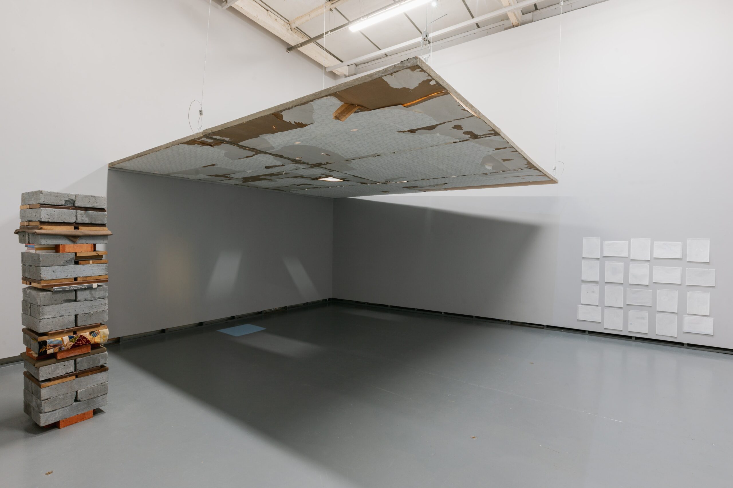 Installation view of an exhibition at Eastside Projects featuring 3 artworks. To the left a monolithic tower of breeze blocks, wood, lace - at the centre a hanging false ceiling with peeling wallpaper - to the right delicate drawings are pinned to the wall in a grid