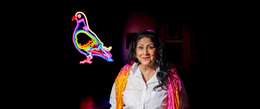 Chila Burman stands next to her artwork: a large drawing of a pigeon made from neon tubing