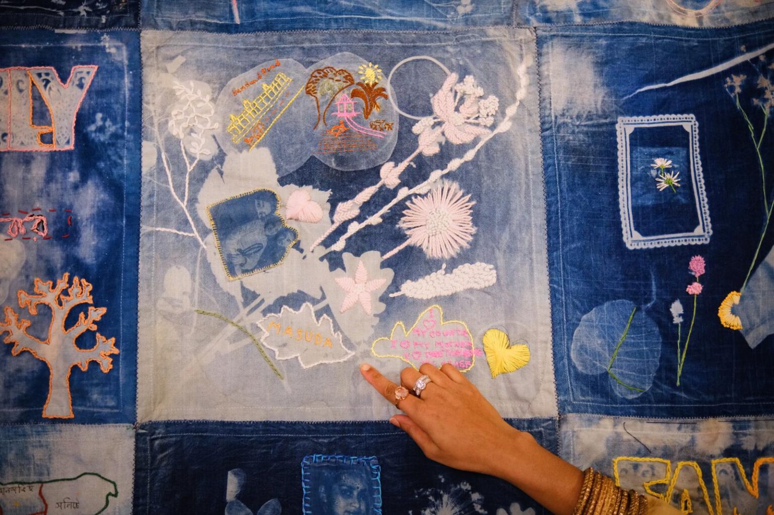 The image is filled with a large patchwork textile made from cyanotypes, in indigo blue, images of leaves and other natural forms in white. Other shaped are embroidered into the cloth with coloured thread. A hand wearing rings, with light brown skin is pointing at an embroidered leaf.