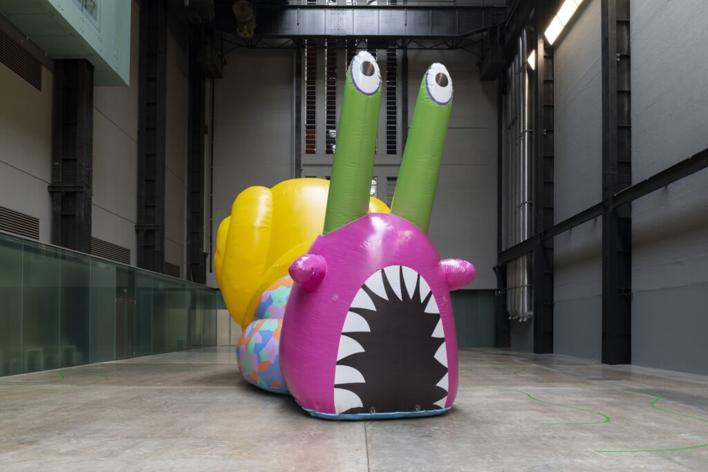 A large inflatable snail sits on the floor inside the cavernous Turbine Hall at Tate Modern. The shell of the snail is bright yellow, the body of the snail is bright pink and the eye stalks are slime green colour. The snail has an open, cartoonish style mouth showing a set of sharp white teeth.
