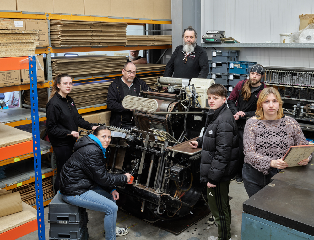 seven white people of varying ages posing around an old industrial looking printing machine