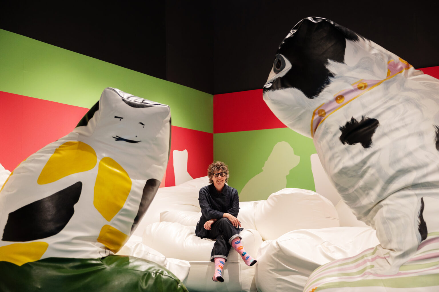 The artist sits on a giant beanbag in a brightly coloured room, flanked by two other giant beanbags shaped like a cat and a dog.