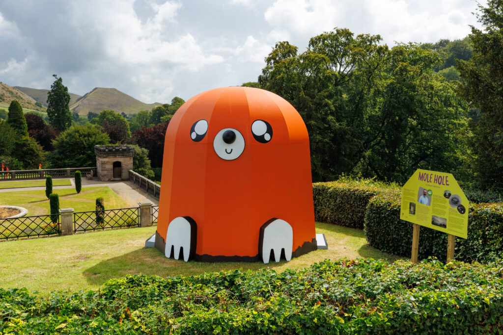 A large orange dome-shaped structure with simplified cartoon facial features resembling a mole emerging from the ground in the garden of a formal English stately home.
