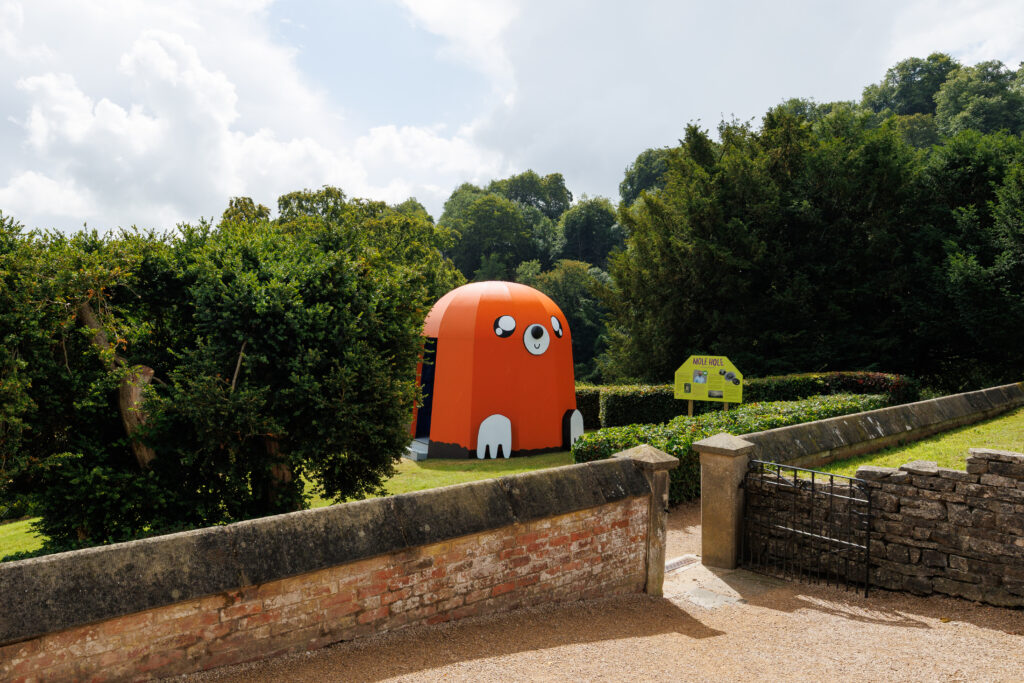 A large orange dome-shaped structure with simplified cartoon facial features resembling a mole emerging from the ground in the garden of a formal English stately home.