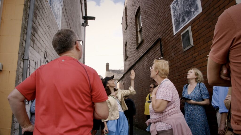 A group of people in a narrow street looking up at a black and white photograph on the wall