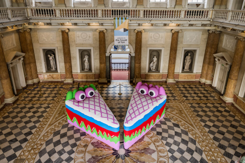 Two large wedge-shaped inflatables sit on the patterned stone floor of an English stately home. They have cartoon eyes and layers of bright colours running through the wedges, resembling slices of cake with bloody teeth.