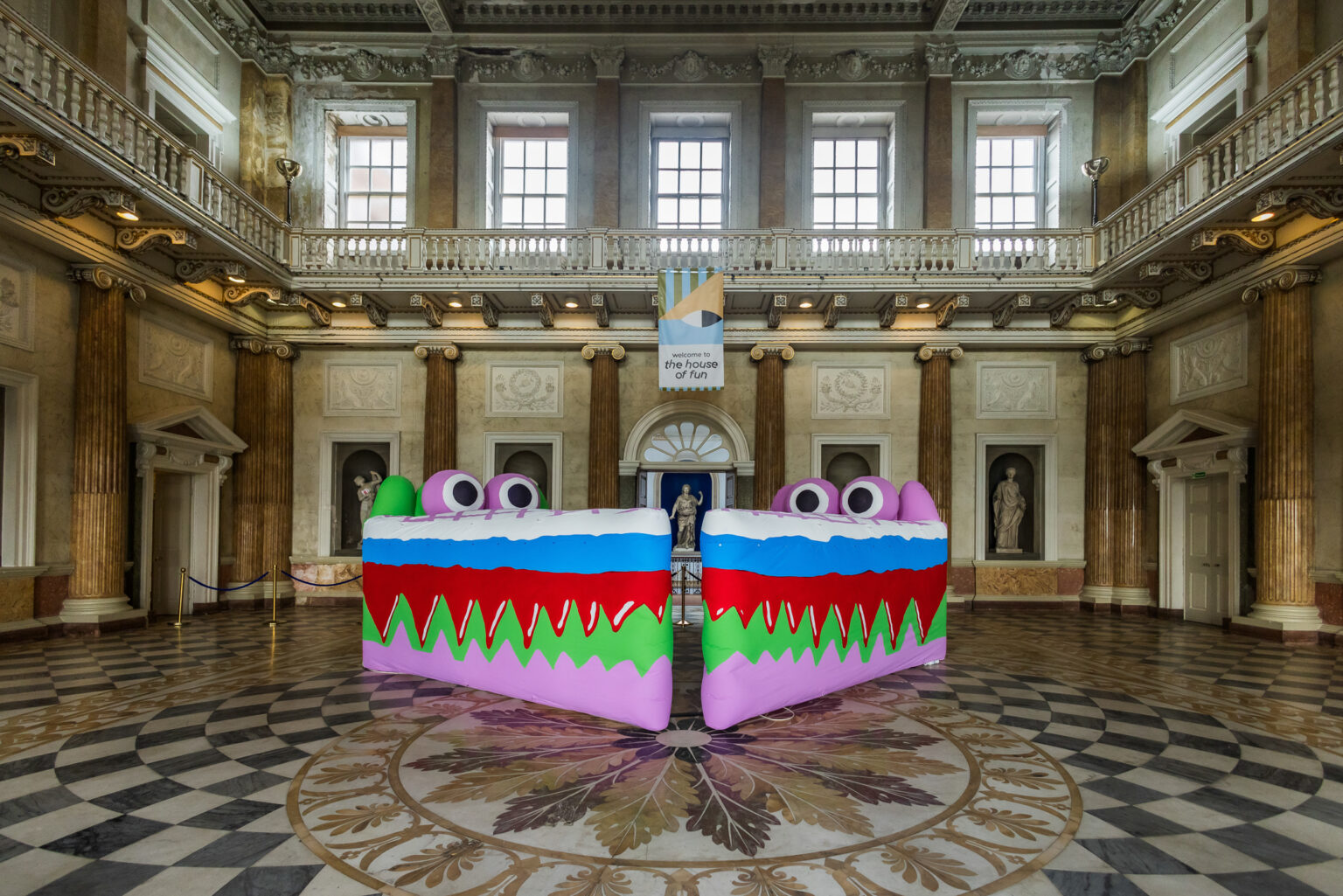 Two large wedge-shaped inflatables sit on the patterned stone floor of an English stately home. They have cartoon eyes and layers of bright colours running through the wedges, resembling slices of cake with bloody teeth.