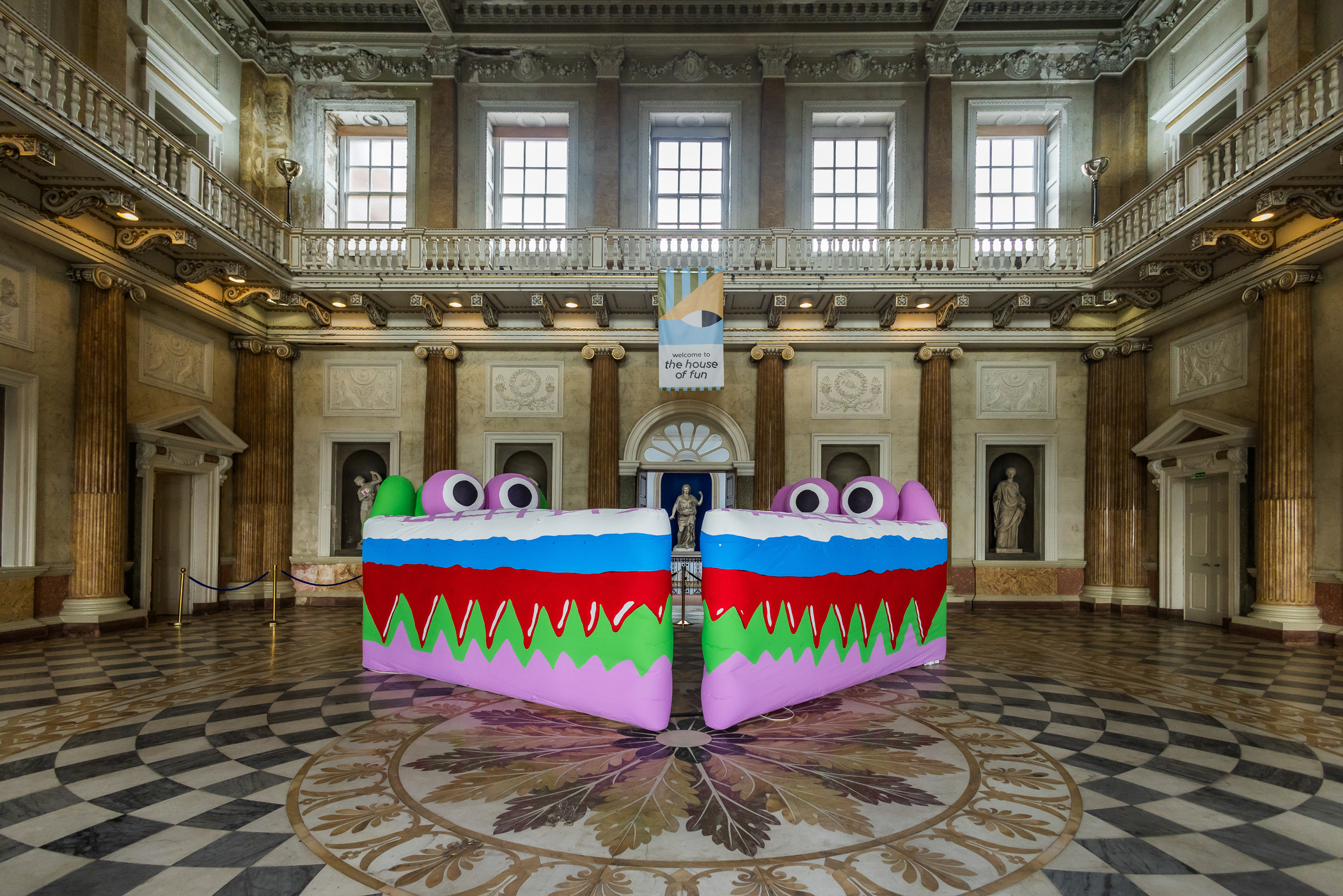 Two large wedge-shaped inflatables sit on the patterned stone floor of an English stately home. They have cartoon eyes and layers of bright colours running through the wedges, resembling slices of cake with bloody teeth.